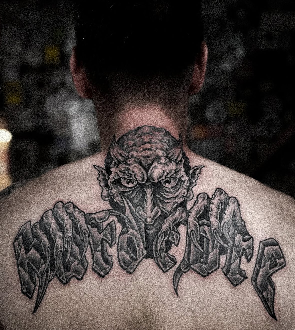 Tattoo of a demonic creature with horns and pointed ears on a person's upper back and neck, with stylized text underneath.