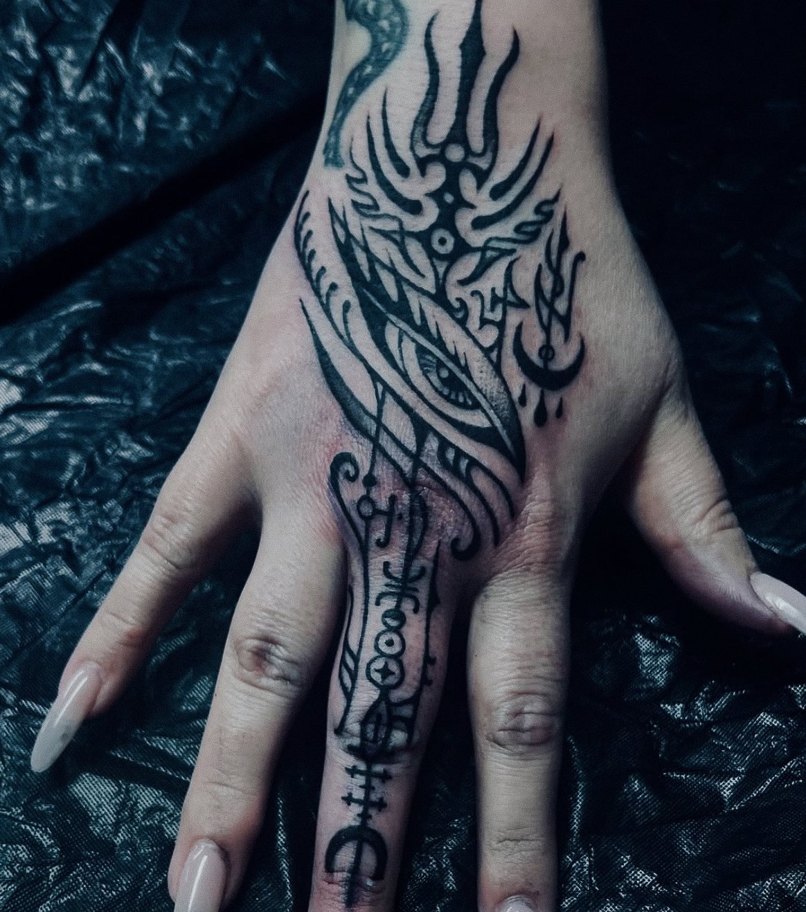 A tattoo on a person's hand and finger featuring abstract and symbolic black ink designs, including an eye, flames, and various symbols.