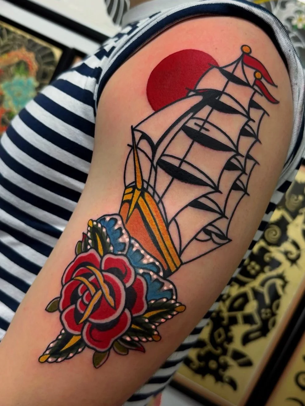 Tattoo of a ship with black outlines, red and yellow details, and a large red sun in the background, along with a red rose with green leaves at the bottom.