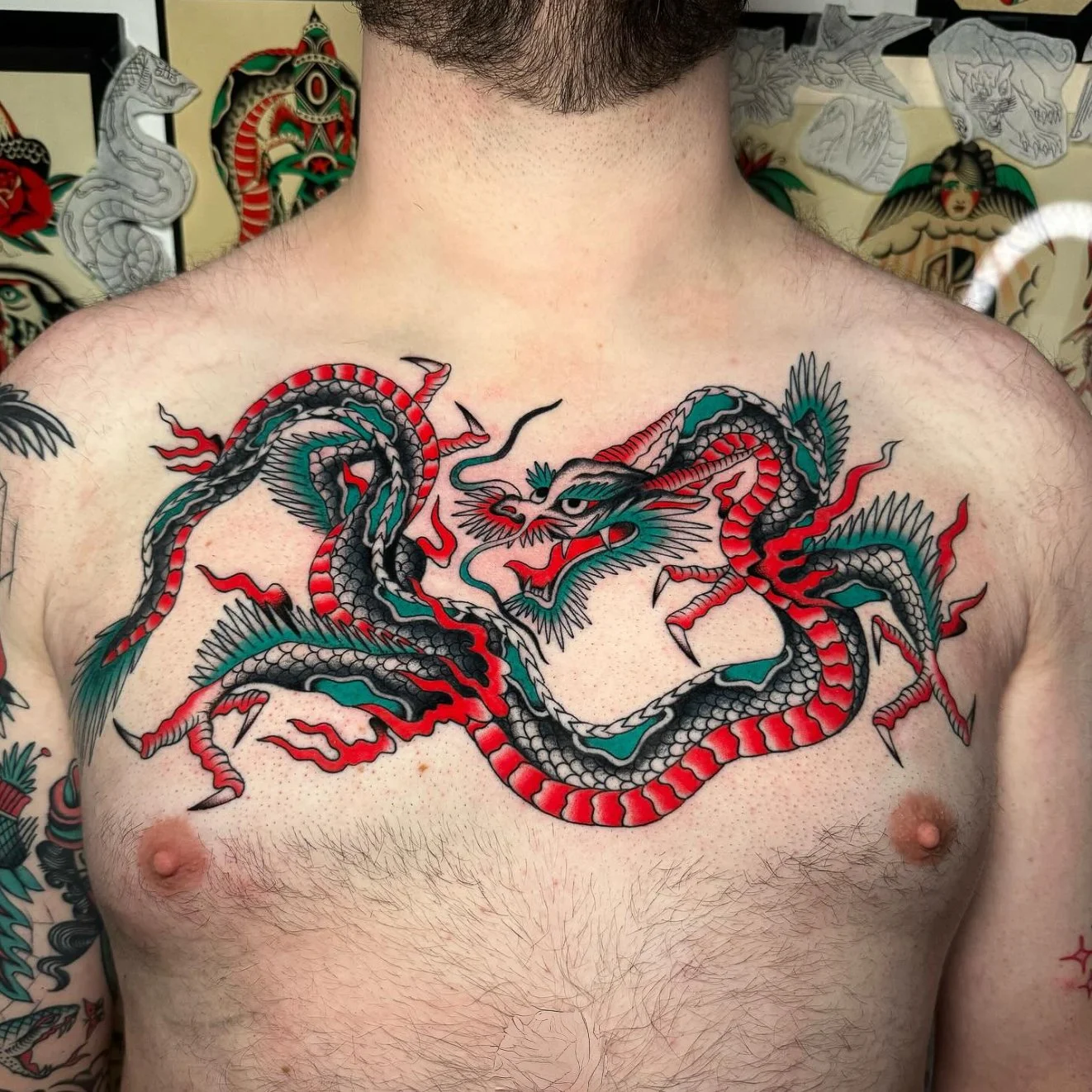 Colorful tattoo of a dragon on a man's chest, featuring intricate details in black, red, teal, and white, with flames and clouds surrounding it.