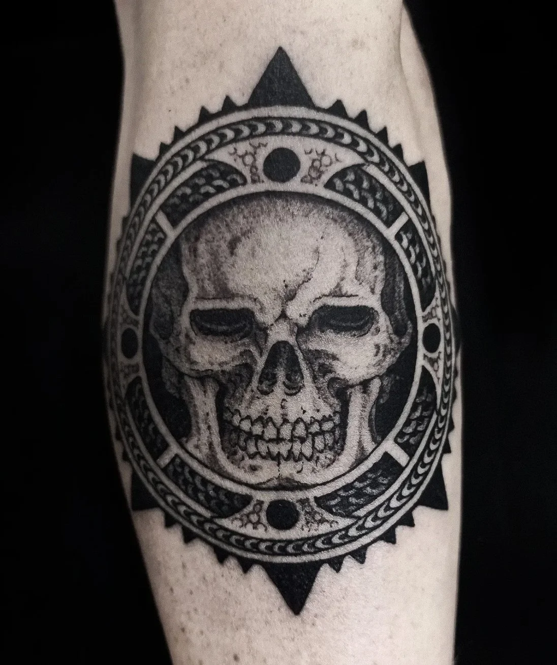 Black and gray skull tattoo surrounded by an ornate circular design with sharp, pointed edges.