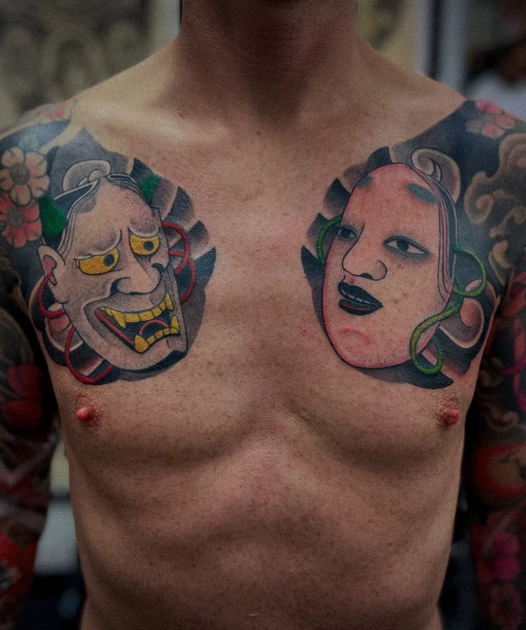 Colorful tattoo of two traditional Japanese masks on a man's chest, one angry and one calm, with floral and decorative elements.