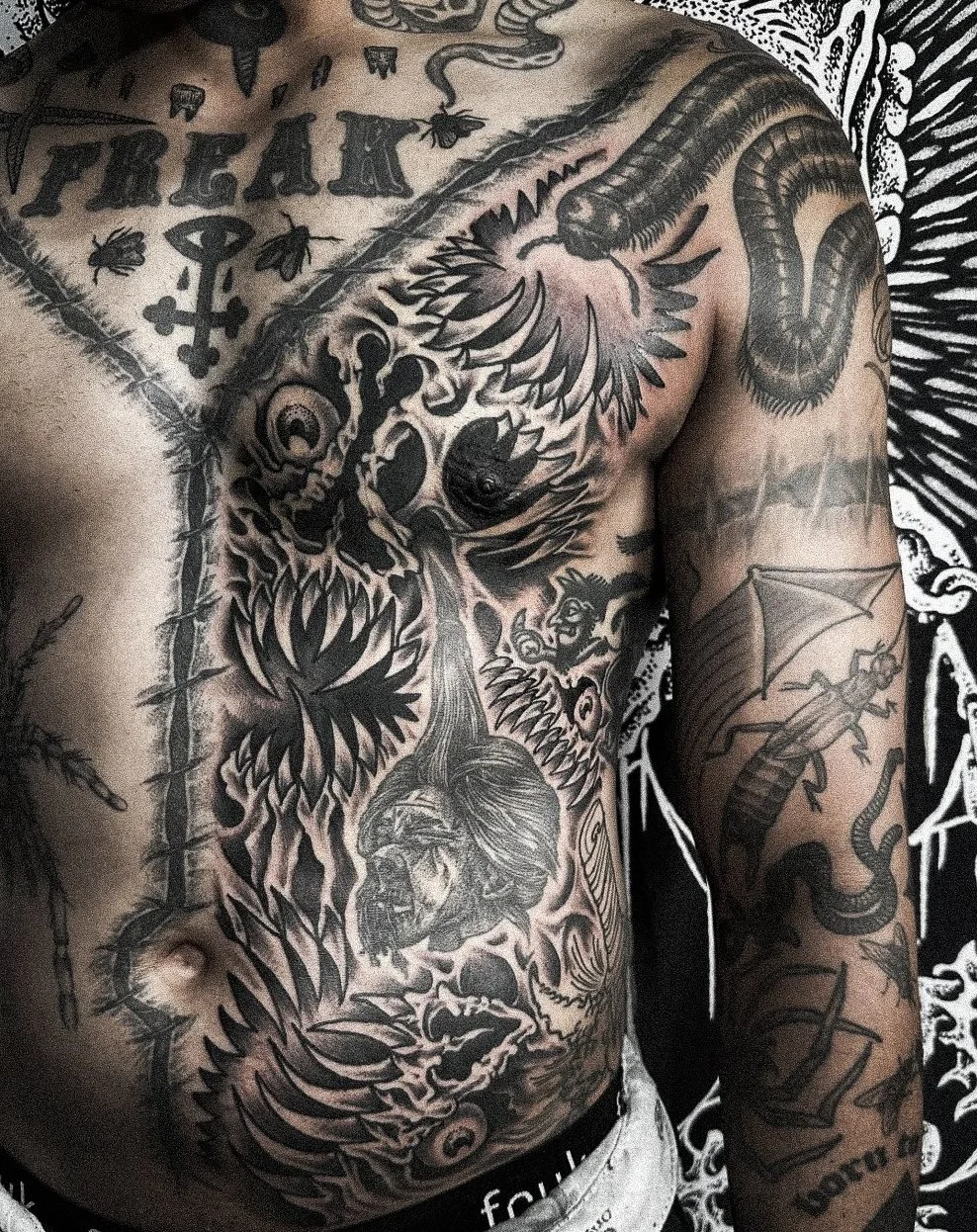 Close-up of a man's upper body showing various black and gray tattoos, including a snake on the shoulder, a skull, a lion, a human face, and a skeleton with wings, with some text and symbols.