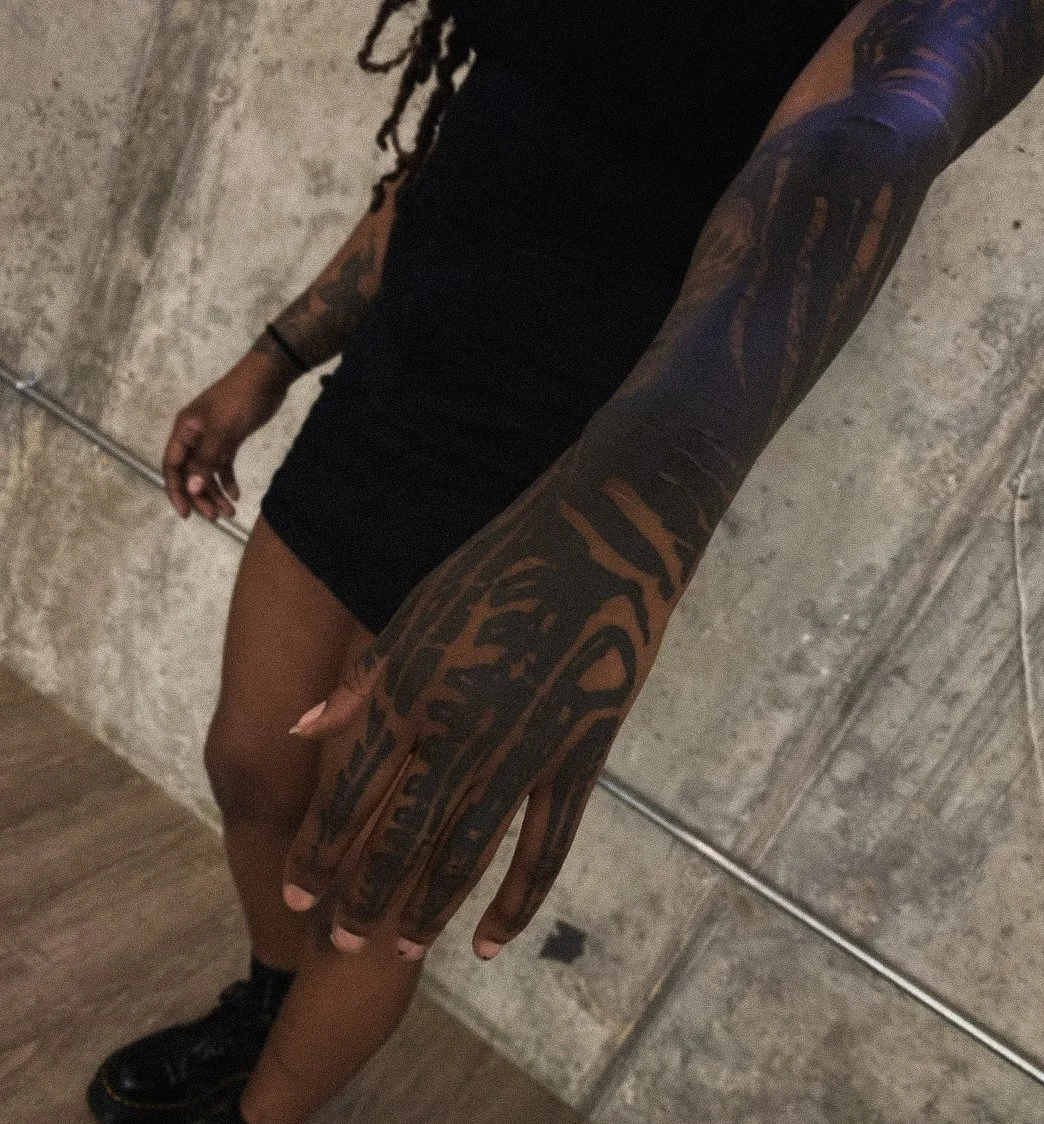 A person with long dreadlocks wearing a black dress and black shoes, showing extensive tattoos on their arm, standing in front of a concrete wall.