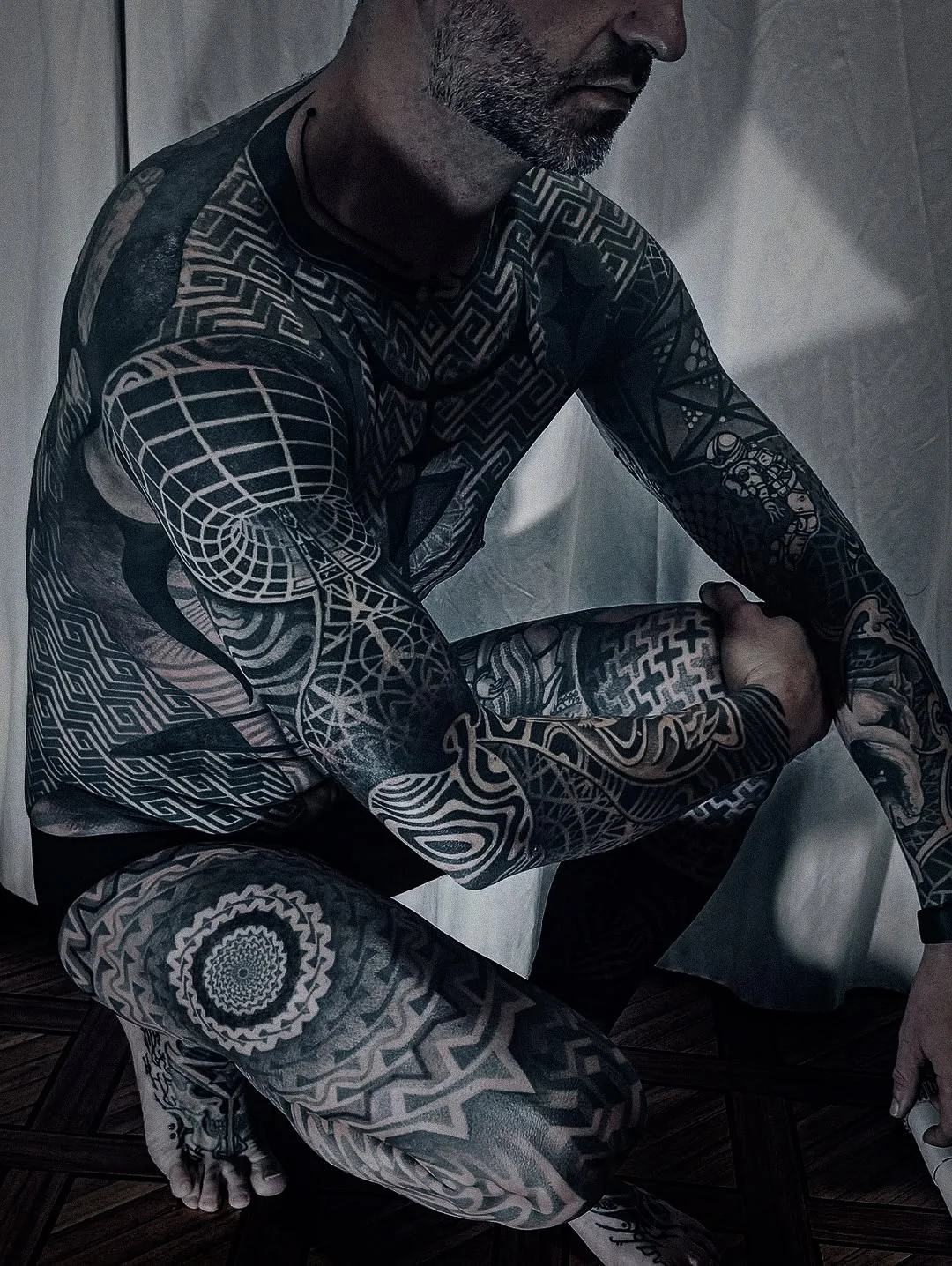 A man with extensive black and white tattoos on his arms, legs, and torso, sitting on a wooden floor with his arms wrapped around one knee, wearing a black shirt, against a plain background.