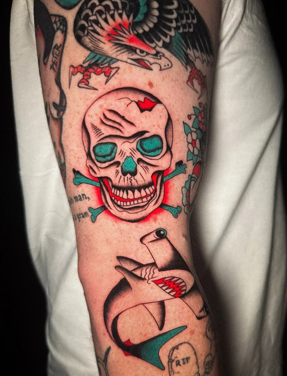 A colorful tattoo featuring a skull with blue eye sockets and a small red heart on its forehead, with green and red accents. Surrounding the skull are various traditional tattoo elements, including a pair of stylized sharks or fish, a bird with outst