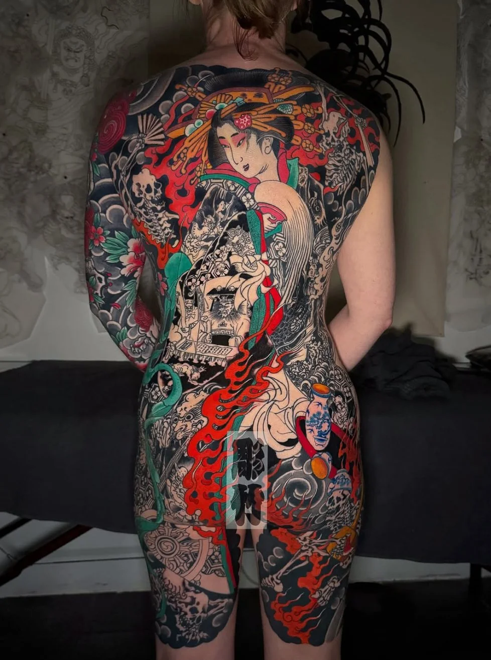 A person with a full back and body tattoo featuring traditional Japanese style art, including a geisha, dragons, skulls, flowers, and waves in vibrant colors.