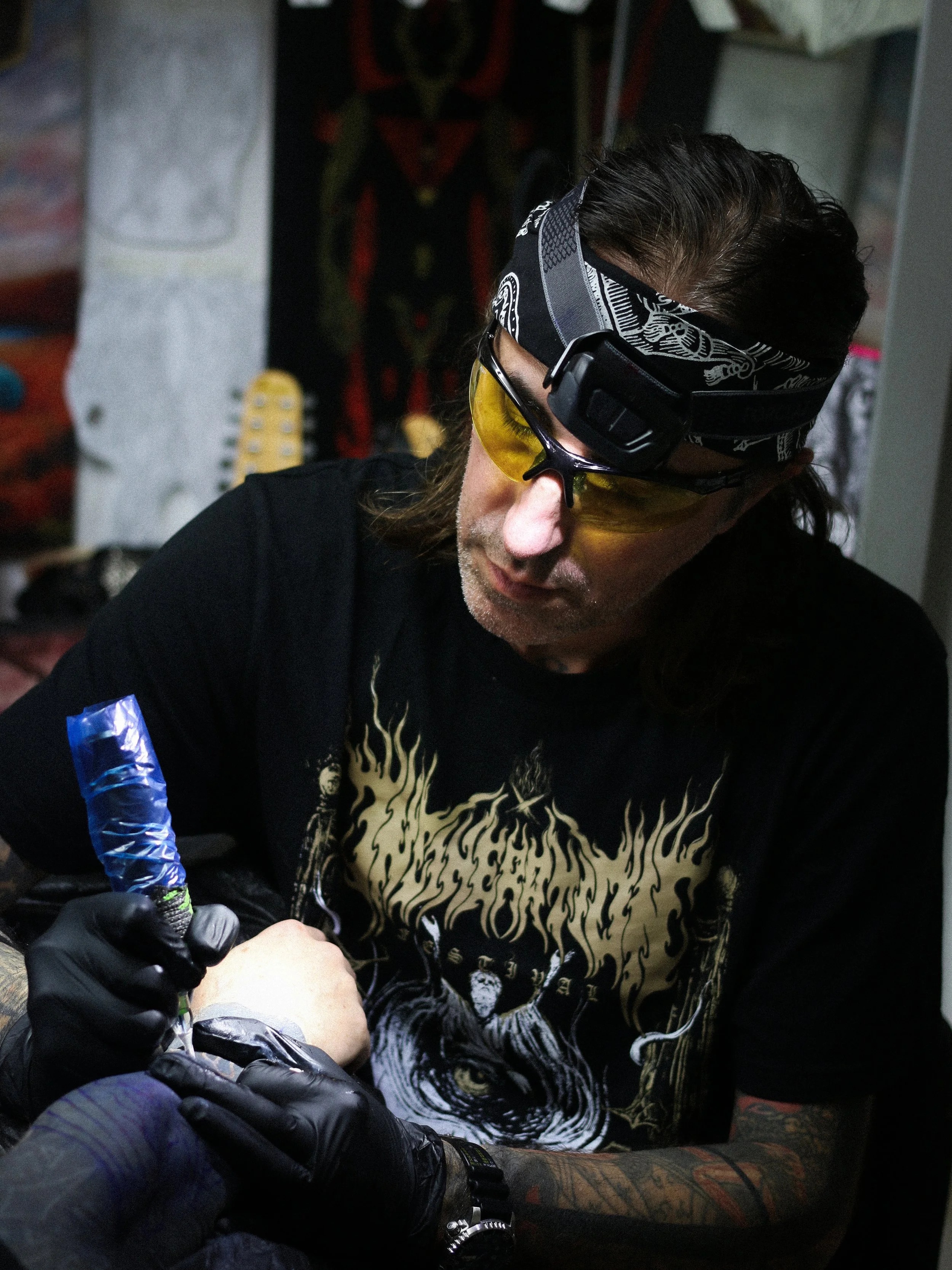 A man with long hair, wearing safety glasses, a headband, and a black t-shirt with a tiger illustration, getting a tattoo on his arm in a tattoo parlor.
