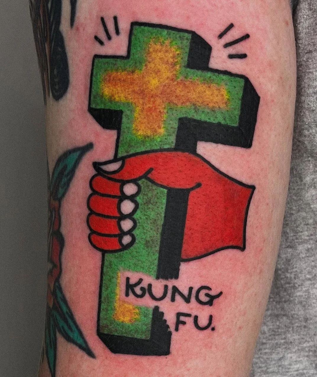 Tattoo of a green cross with yellow and orange shading, held by a red hand with black outline, with the words 'KUNG FU.' written below.