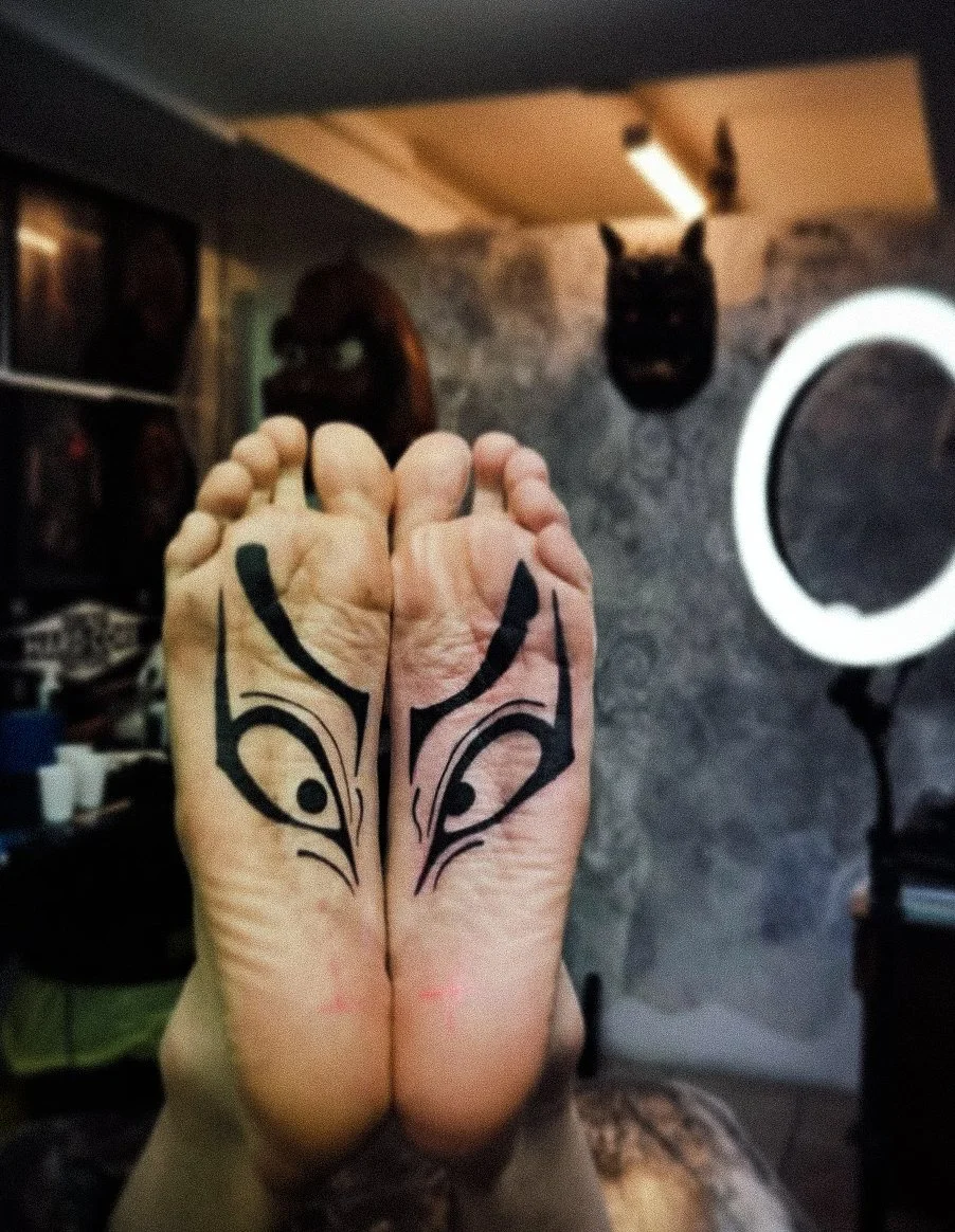 A person’s feet with tribal-style black ink tattoos on the tops of both feet. In the background, a room with dark walls, photographs, a mounted animal head, and a ring light are visible.