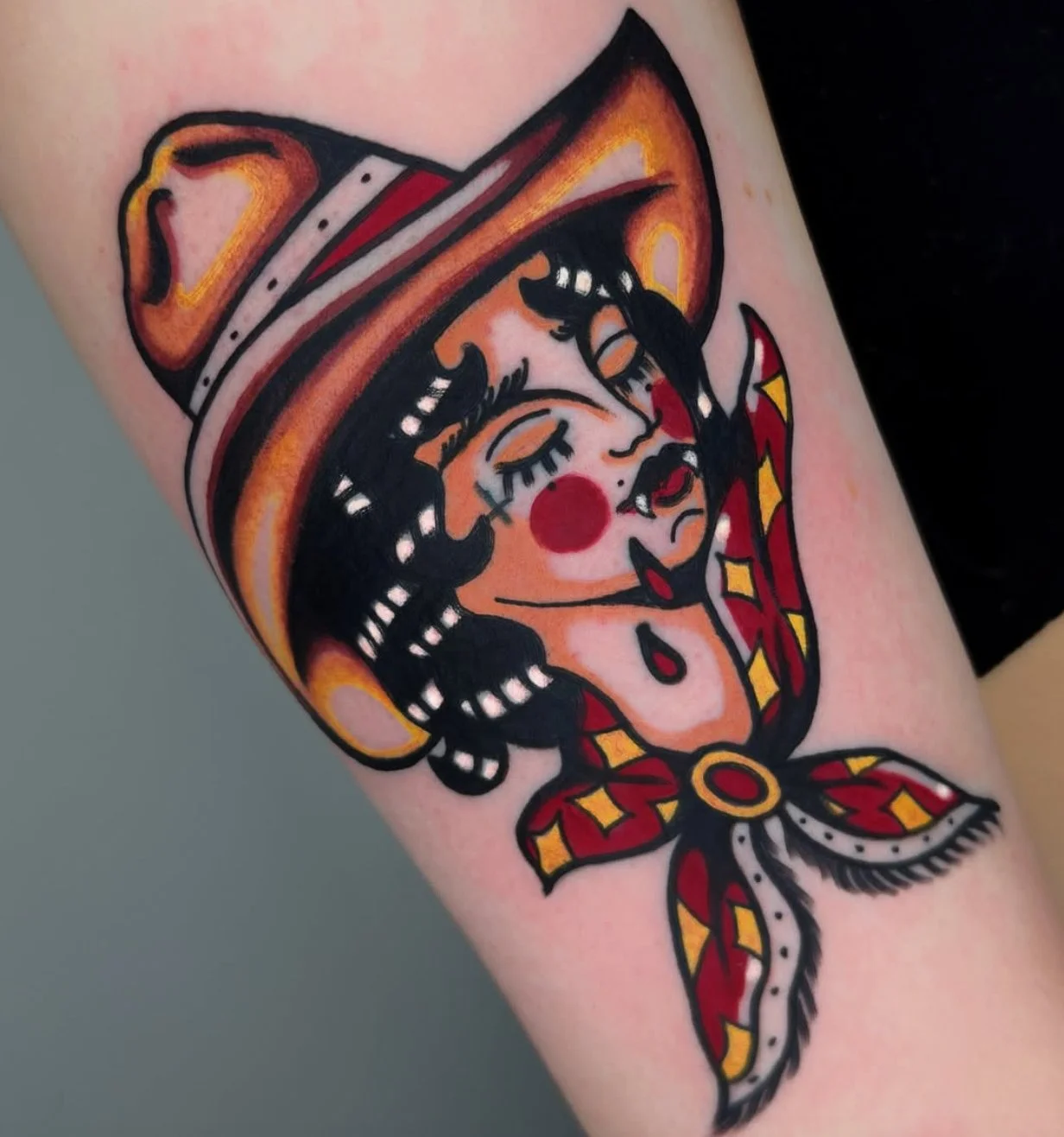 Tattoo of a woman with clown makeup, wearing a cowboy hat with a red band, and a red and yellow scarf around her neck.