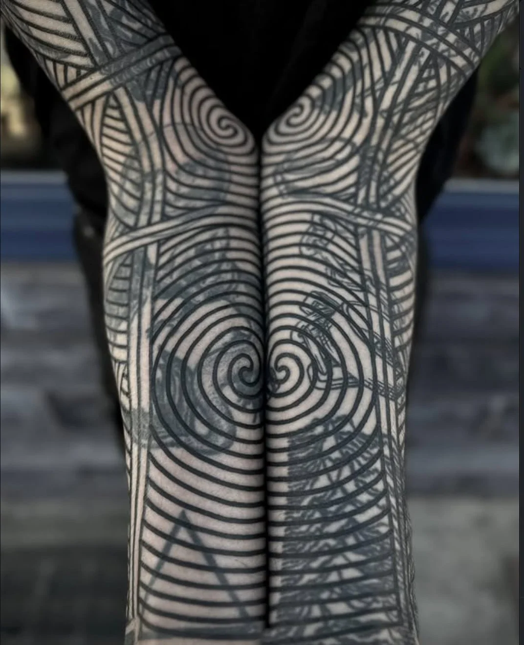 Person's legs with black geometric spiral and line tattoos, crossed at the ankles, standing on a sidewalk.