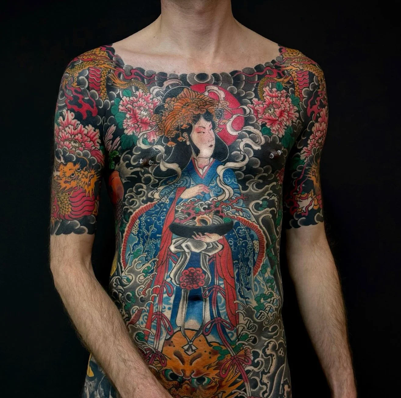 A person with extensive traditional Japanese-style body tattoos featuring a woman in a kimono among clouds, cherry blossoms, dragons, and a tiger, set against a black background.
