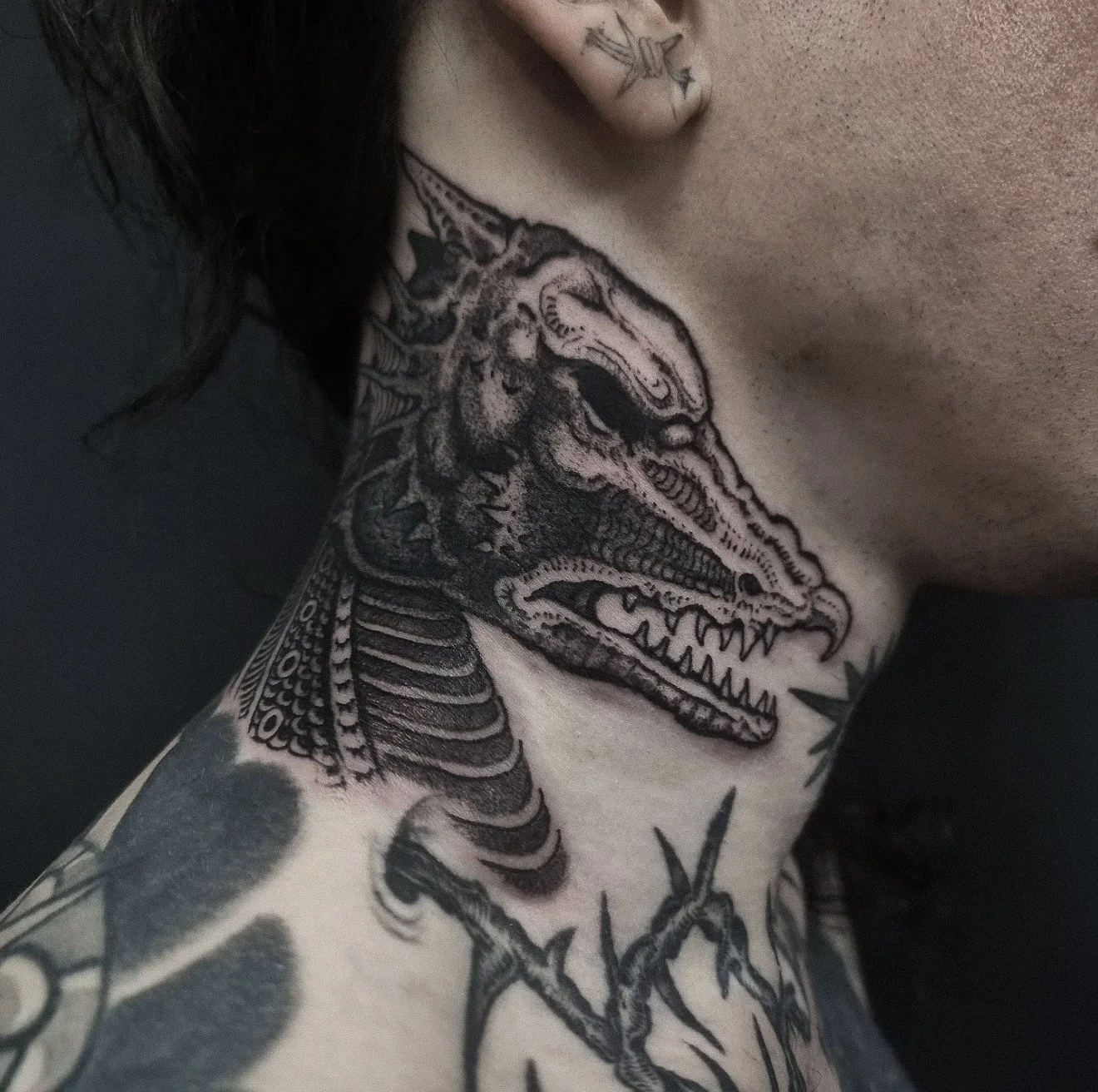 Close-up of a person's chest and shoulder tattoo featuring a detailed dinosaur, possibly a Tyrannosaurus rex, with its mouth open and teeth showing. Additional tattoos are visible on the person's neck and arm.