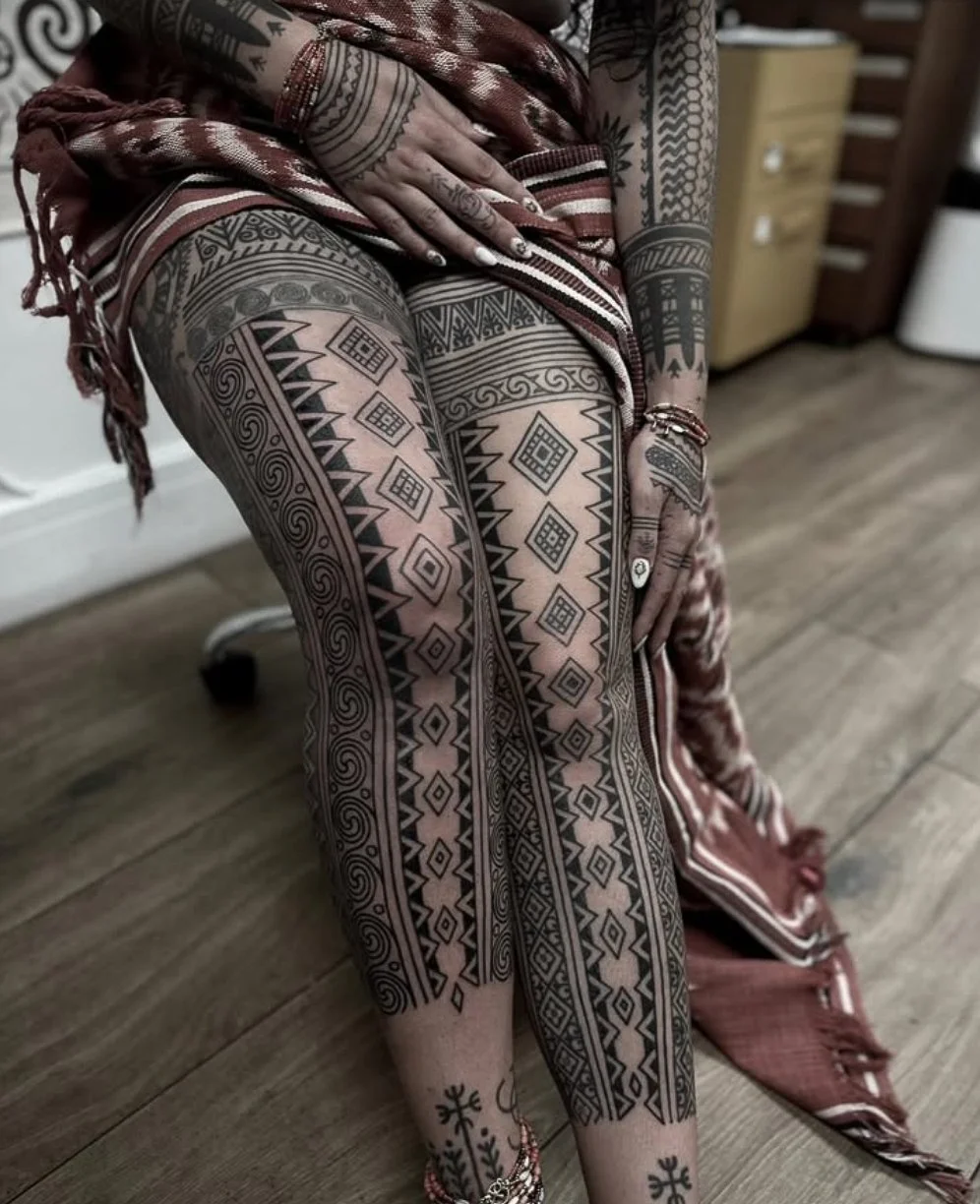 A woman with extensive tribal tattoos on her legs, arms, hands, and fingers, sitting on a chair. She is wearing a patterned dress, and her nails are decorated with designs. The background includes a wooden floor and furniture in a room.