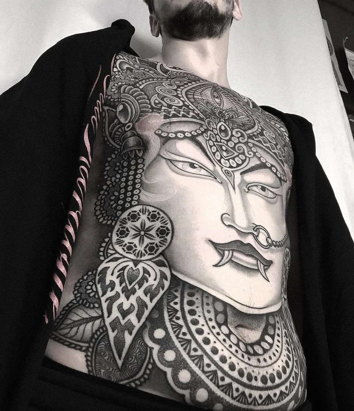 Black and white tattoo of a woman's face with traditional ornamentation, aligned with Asian cultural elements, covering the person's chest and shoulder.