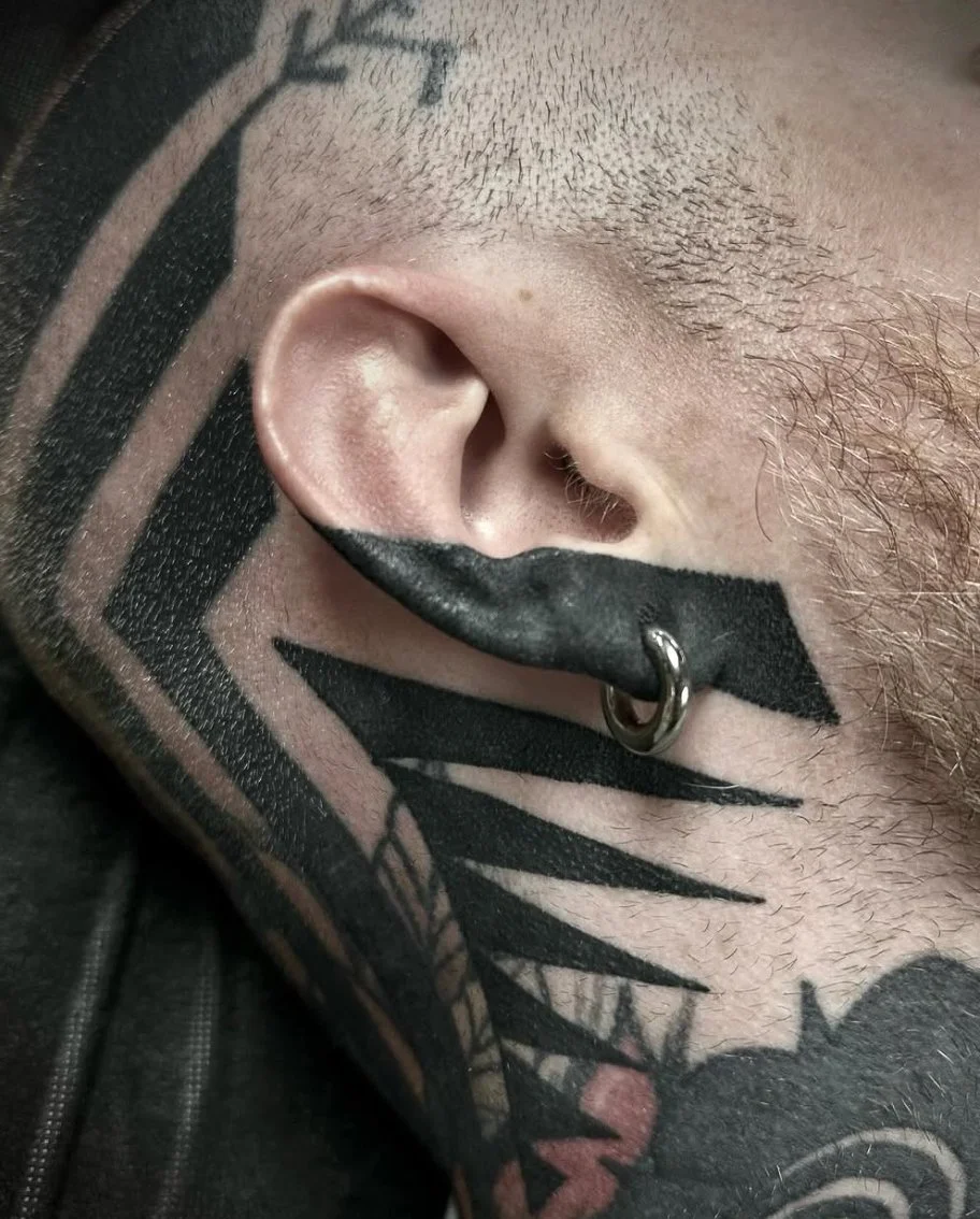 Close-up of a person's face showing an ear, part of a tattooed neck, and facial hair. The person has a hoop earring and ties or ribbons piercing through the lip.