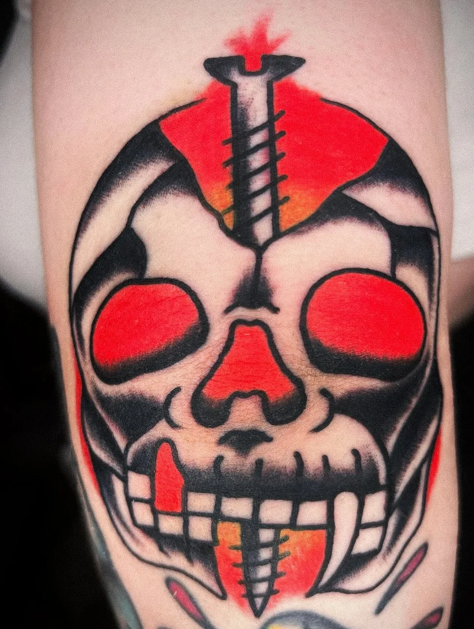 Colorful tattoo of a stylized skull with a large syringe crossing through the top, with red and yellow shading behind it.