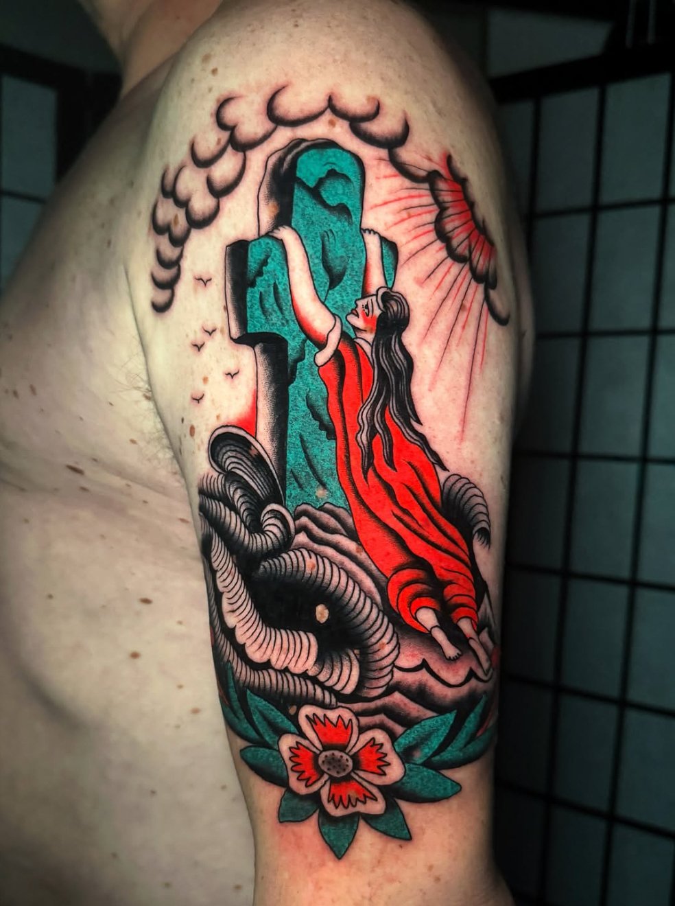 Tattoo of a woman with long black hair in a red dress. She is reaching up towards a blue-faced statue with a long nose. The scene includes steam or clouds, a flower at the bottom, and a red sun with rays in the background.