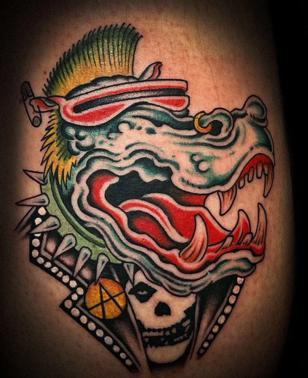 Tattoo of a colorful dragon's head with sharp teeth and a skull at the base, decorated with a pentagram symbol.