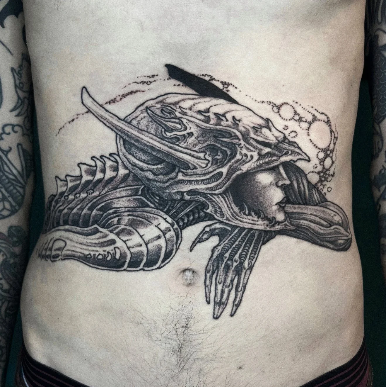 Black and gray detailed tattoo on person's chest featuring a surreal scene with a person with eyes closed, with an animal skull, and various abstract and organic shapes.