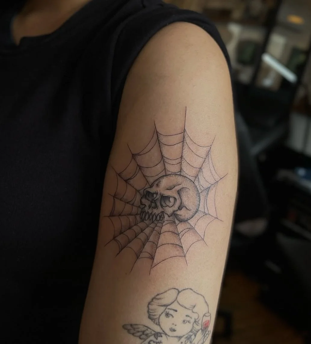Tattoo of a skull with a crescent moon in a spider web design on the person's upper arm.