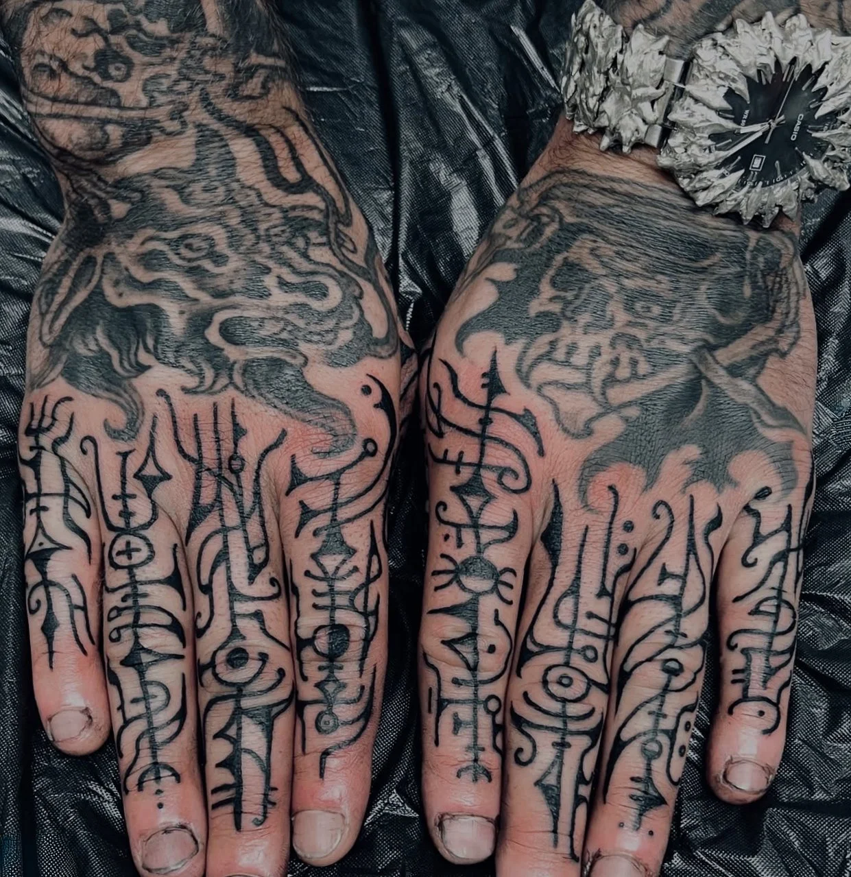 Close-up of two tattooed hands with black and gray gothic and mythological-themed tattoos, lying on a dark fabric surface. The tattoos include faces, creatures, and intricate symbols.