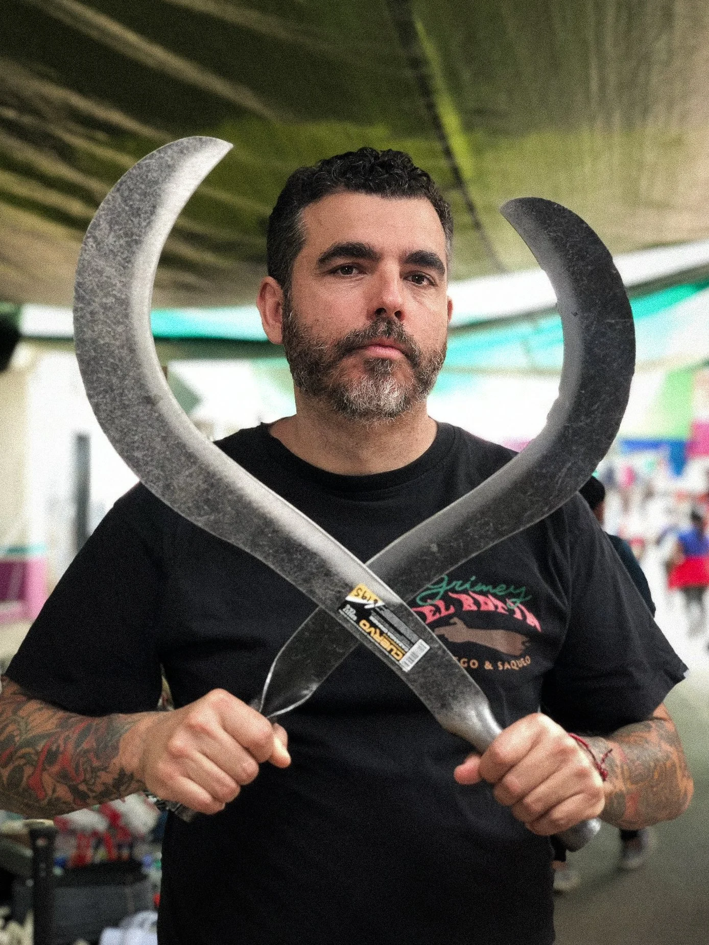 A man with a beard and tattoos holding two curved blades crossed in front of him at a marketplace or indoor event.