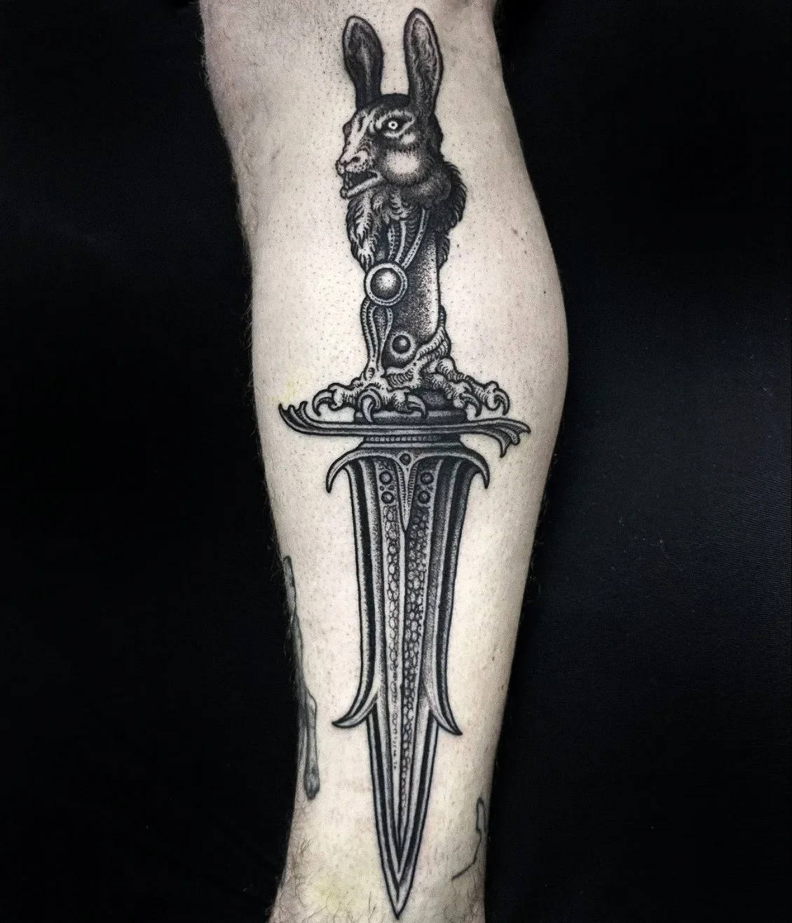 Tattoo of a sword with a rabbit's head and a beard, featuring intricate linework and shading.