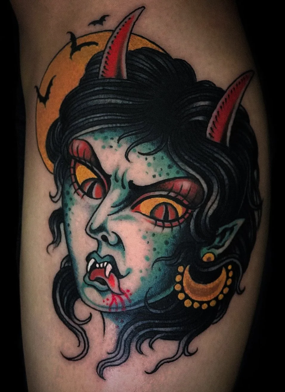 Tattoo of a mythical creature with green skin, red eyes with black slits, black hair, purple horns, and fanged mouth, set against an orange moon with bats flying around.