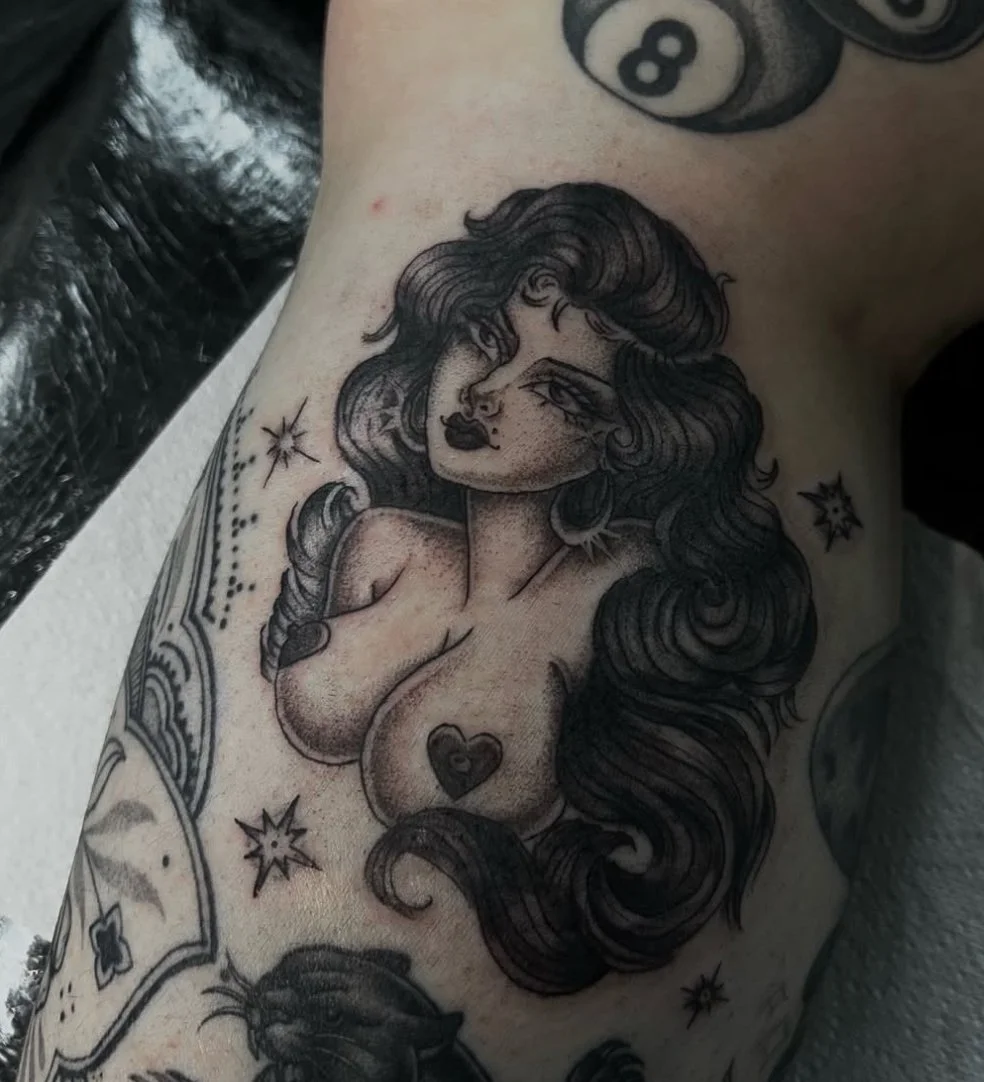 Black ink tattoo of a woman with long wavy hair, bold makeup, and large hoop earrings, surrounded by stars and decorative elements.