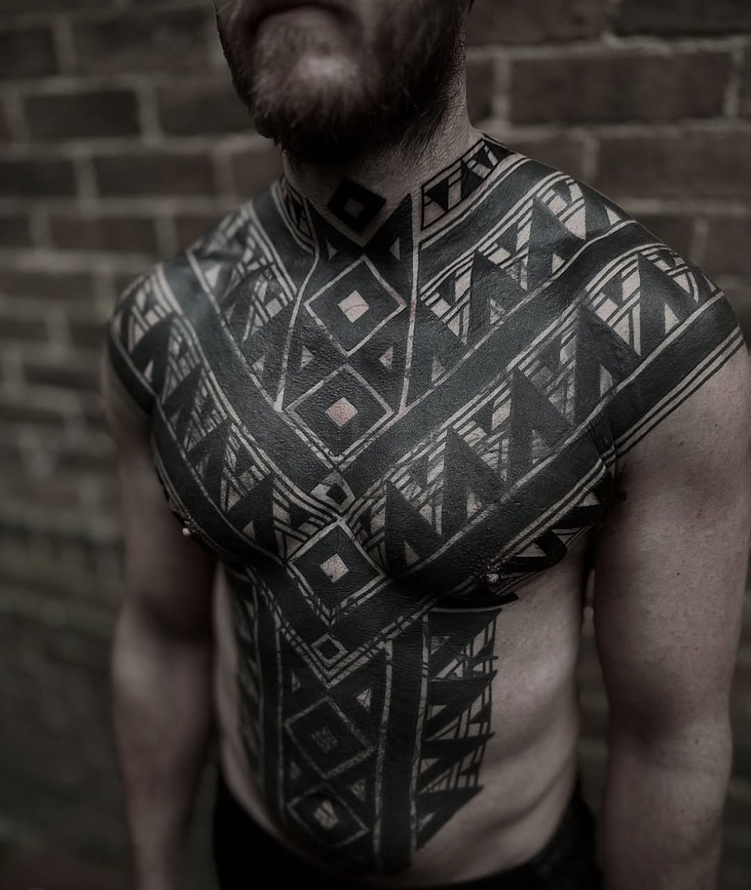 A man with a beard and mustache has an intricate black geometric tattoo covering his chest, shoulders, and neck, set against a brick wall background.