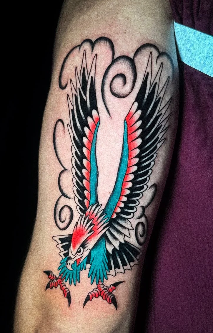 Tattoo of a colorful eagle with wings spread wide, featuring black, red, and blue details, surrounded by black and gray clouds on someone's arm.