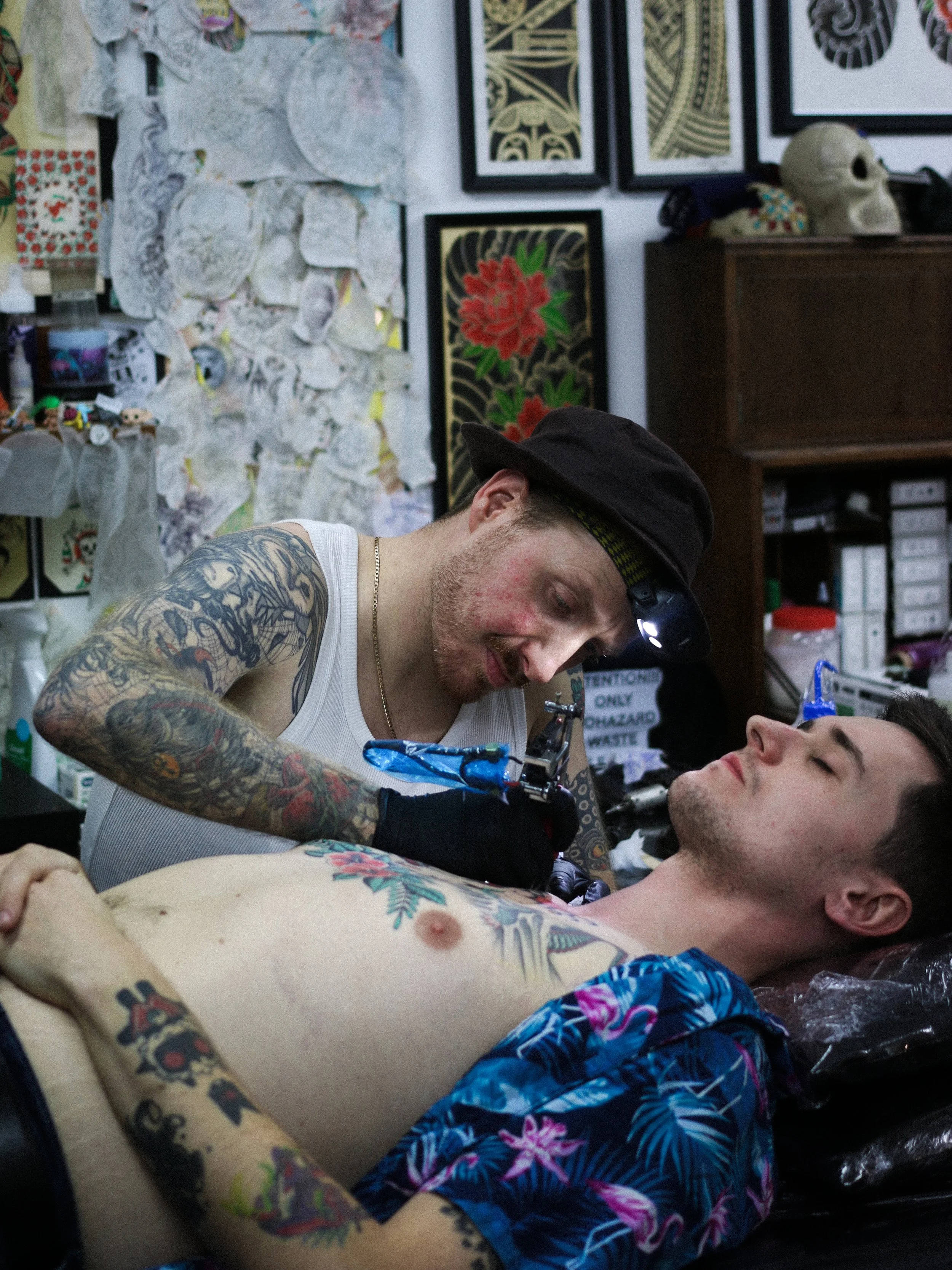A tattoo artist in a white tank top and a black hat tattoos a man with short dark hair who is lying shirtless on a tattoo table, in a room decorated with framed art and tattoo designs.