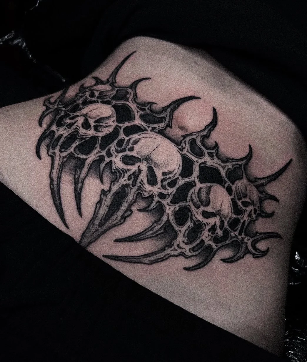 Black and gray tattoo of three skulls with intricate, spiked, and flame-like designs on skin.