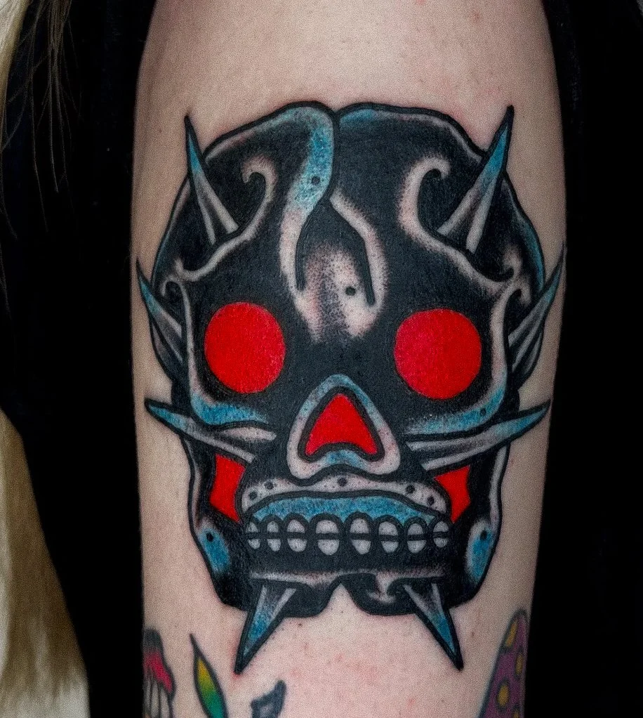 Tattoo of a stylized skull with glowing red eyes and pointed spikes, in dark colors with blue accents.