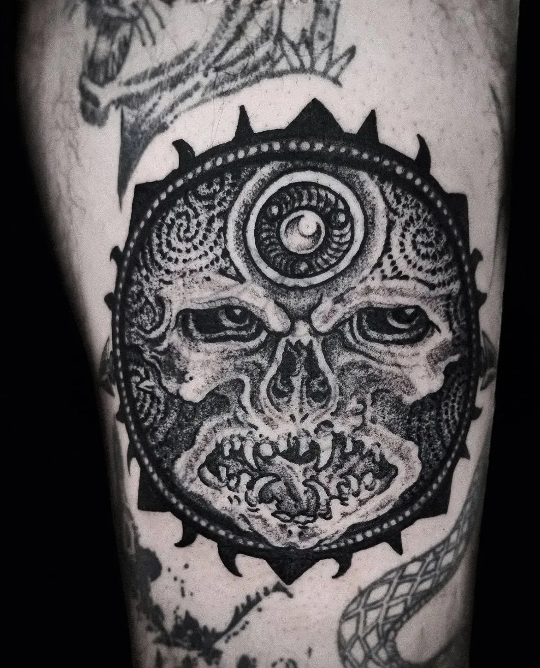 A tattoo of a skull with intense eyes, surrounded by intricate patterns and a circular design with a small crescent moon and dotted details on the forehead.