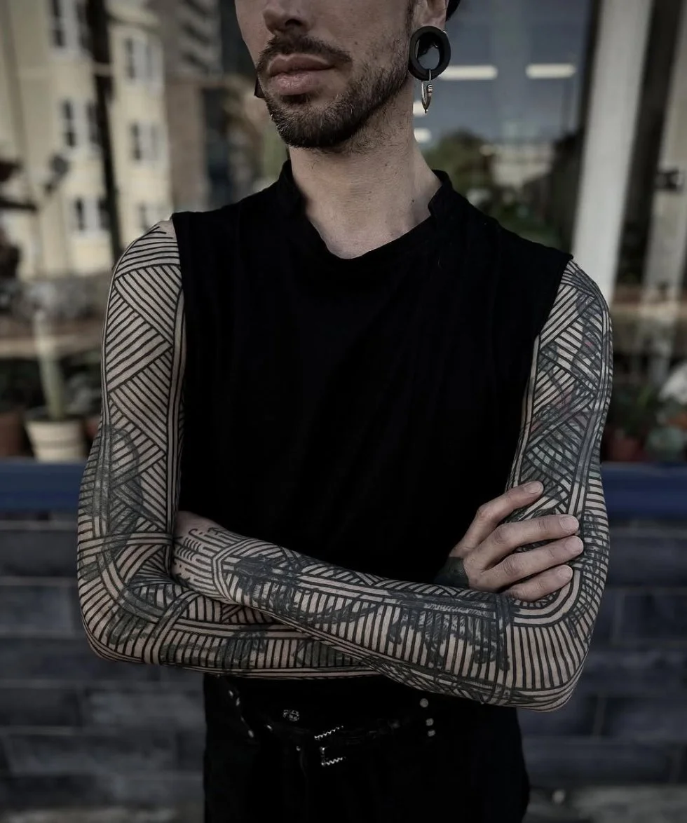 A man with tattoos on both arms, wearing a sleeveless black shirt and a large gauge earring, crossing his arms outdoors.