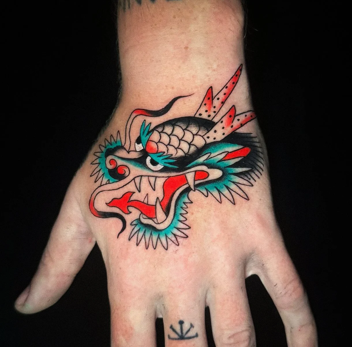 A colorful dragon tattoo on a person's hand, featuring a fierce face with sharp teeth, whiskers, and red and black accents, with a black background.