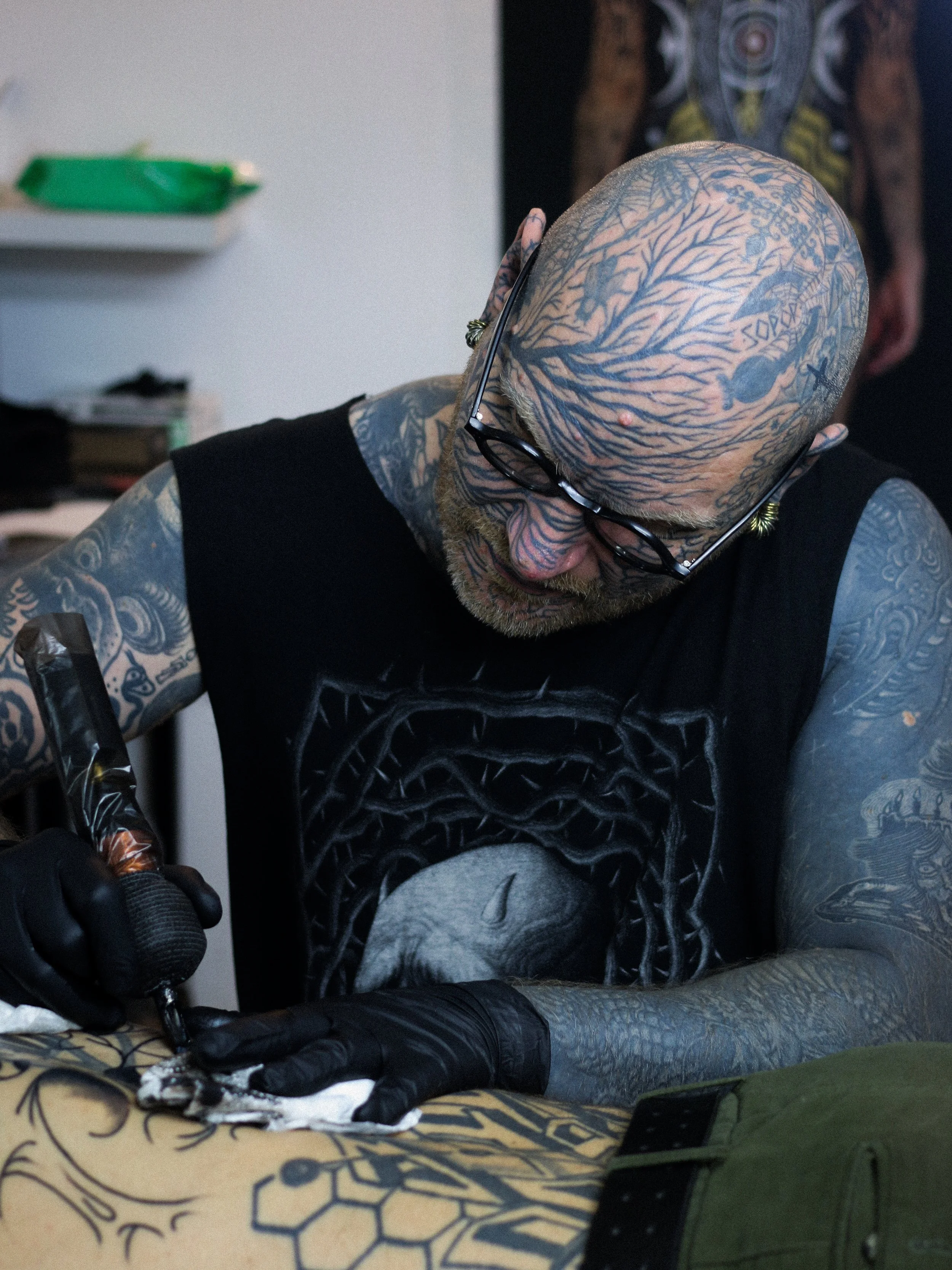 A bald, heavily tattooed man wearing glasses and black gloves, tattooing in a room.