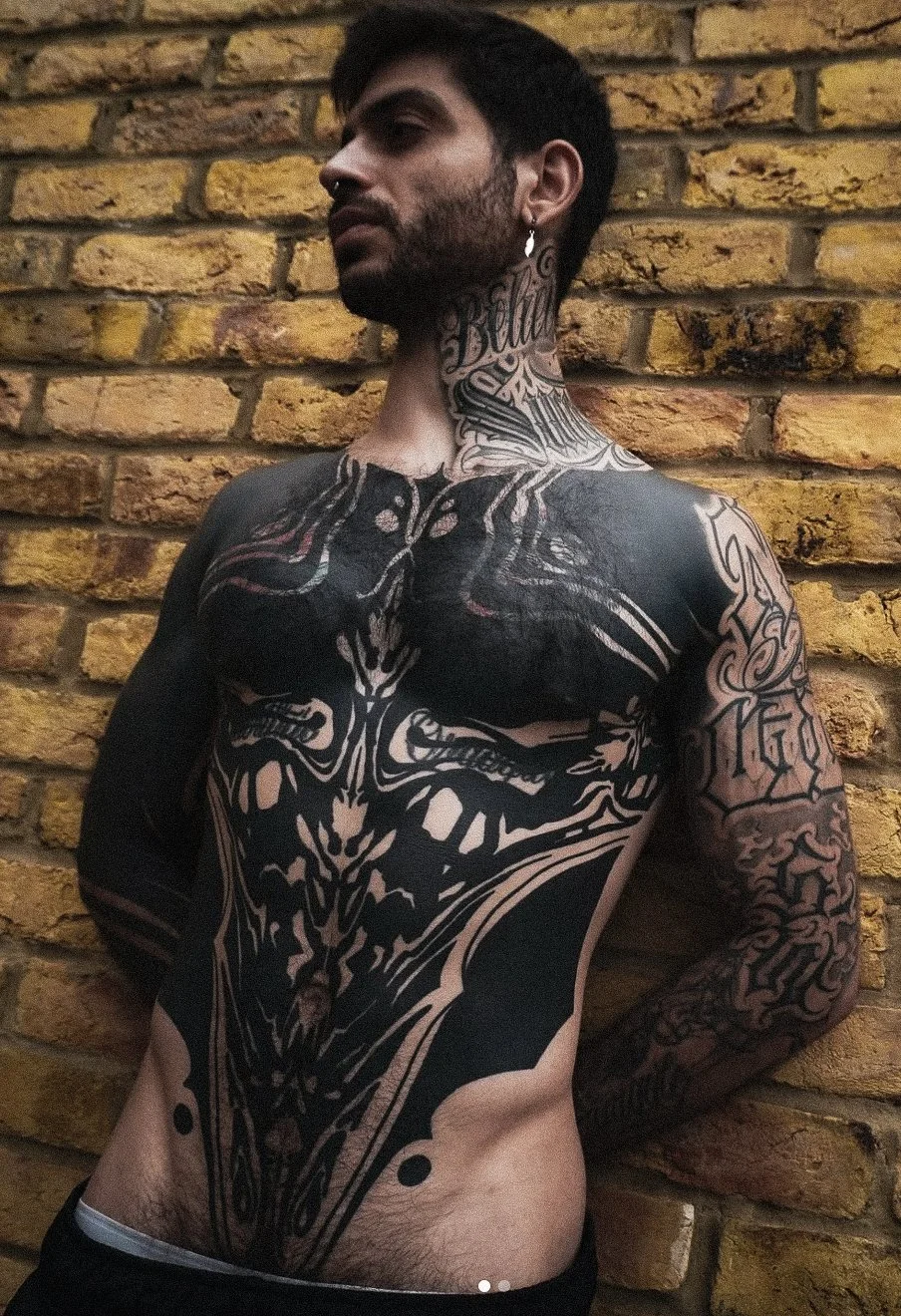 A man with dark hair, facial hair, and tattoos on his neck, chest, and arms, standing against a yellow brick wall. He has a septum piercing and wears an earring.