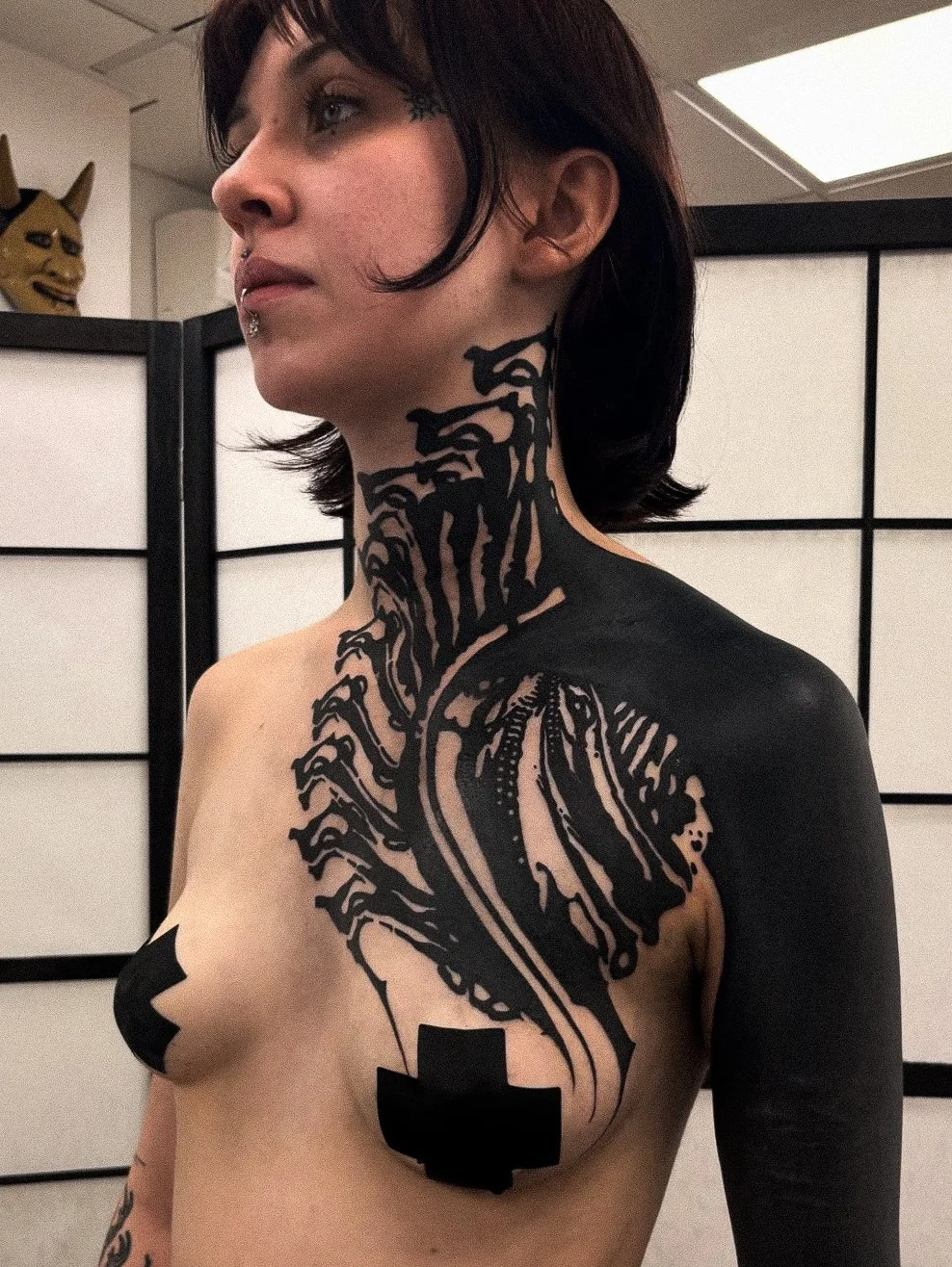 A woman with dark, chin-length hair and tattoos on her neck, chest, and shoulder. She has black body paint or ink art covering part of her neck, shoulder, and chest, with a large tattoo of a fist on her breast and a black cross on her upper arm. In t
