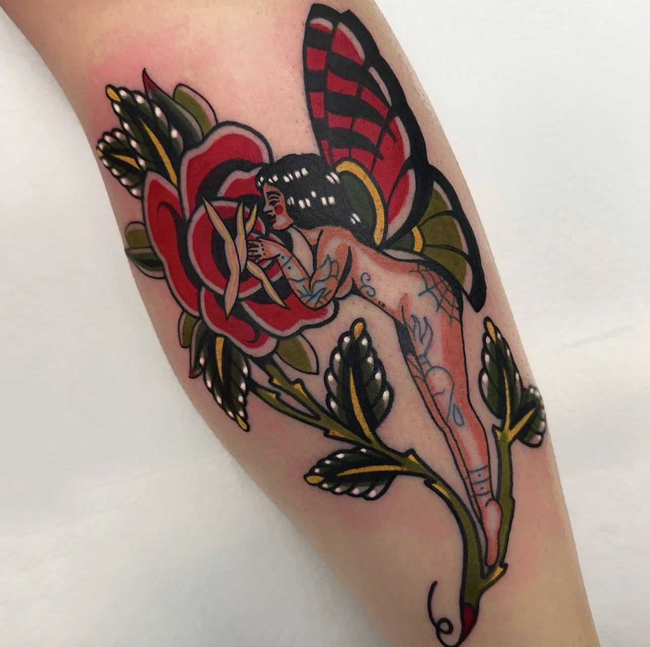 Tattoo of a fairy with butterfly wings, sitting on a flower, with a dragonfly perched on her hand, surrounded by leaves.