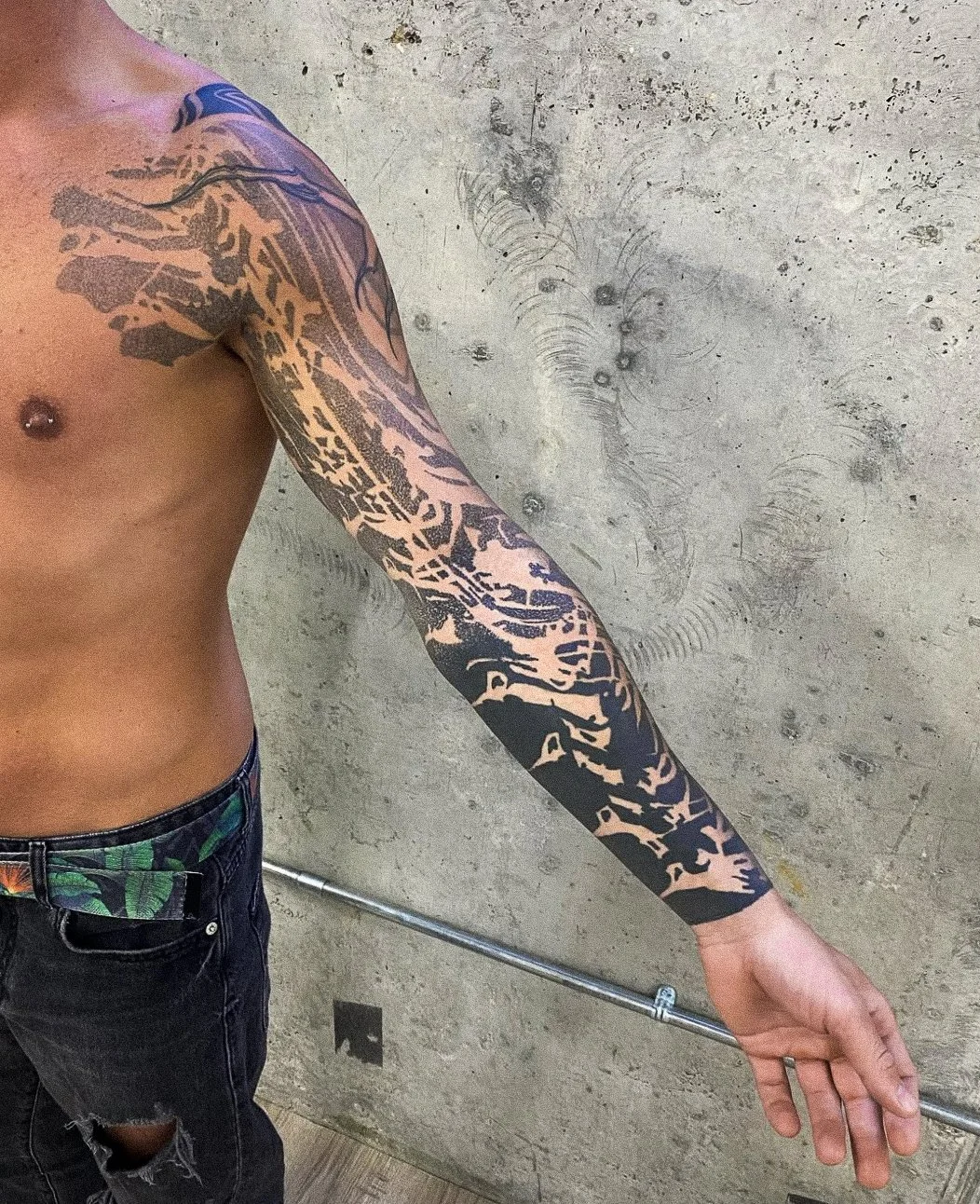 A person with a geometric black and beige tattoo sleeve on their arm, wearing black pants with a colorful belt, standing against a concrete wall.