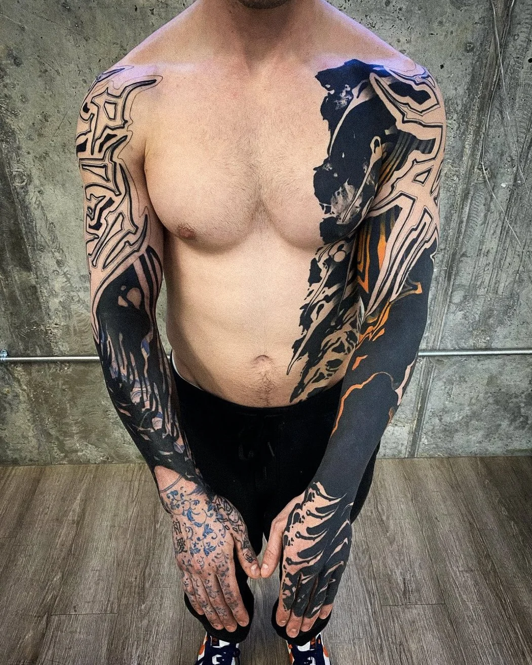 A shirtless man with extensive black and orange tattoos on his arms, chest, and hands, standing in front of a concrete wall.