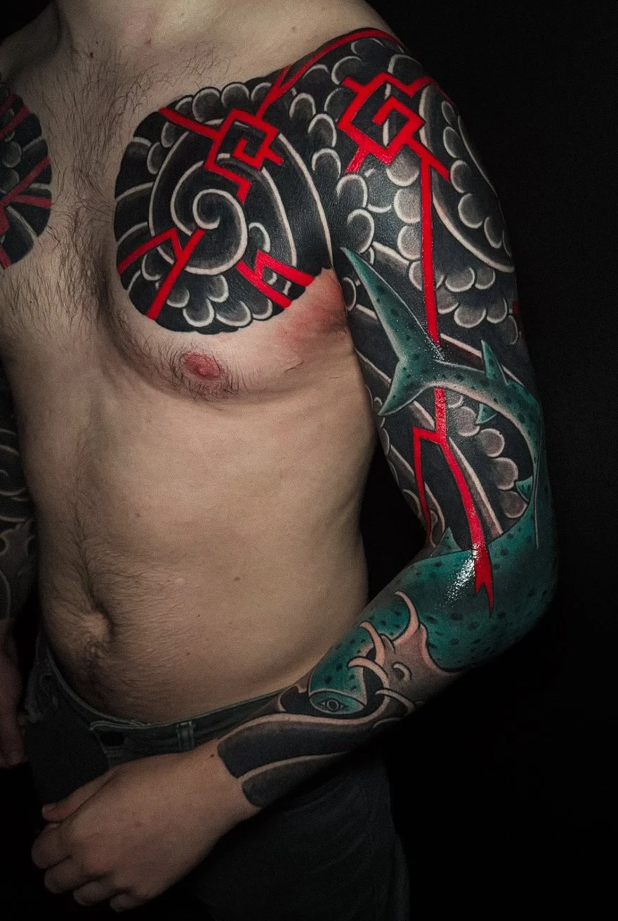 A person with a large colorful tattoo covering the shoulder, upper arm, and part of the chest. The tattoo features a stylized shark, red geometric lines, and swirling clouds in black, gray, and teal colors.