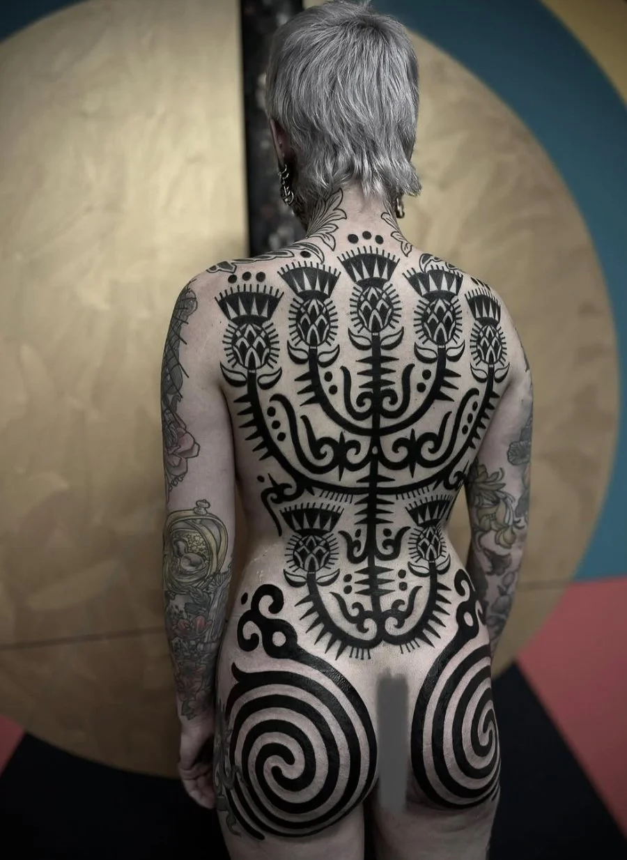 Back view of a person with gray hair and multiple tattoos, including large black tribal-style tattoos on their back and smaller tattoos on their arms, standing in front of a colorful background with gold, blue, and pink sections.