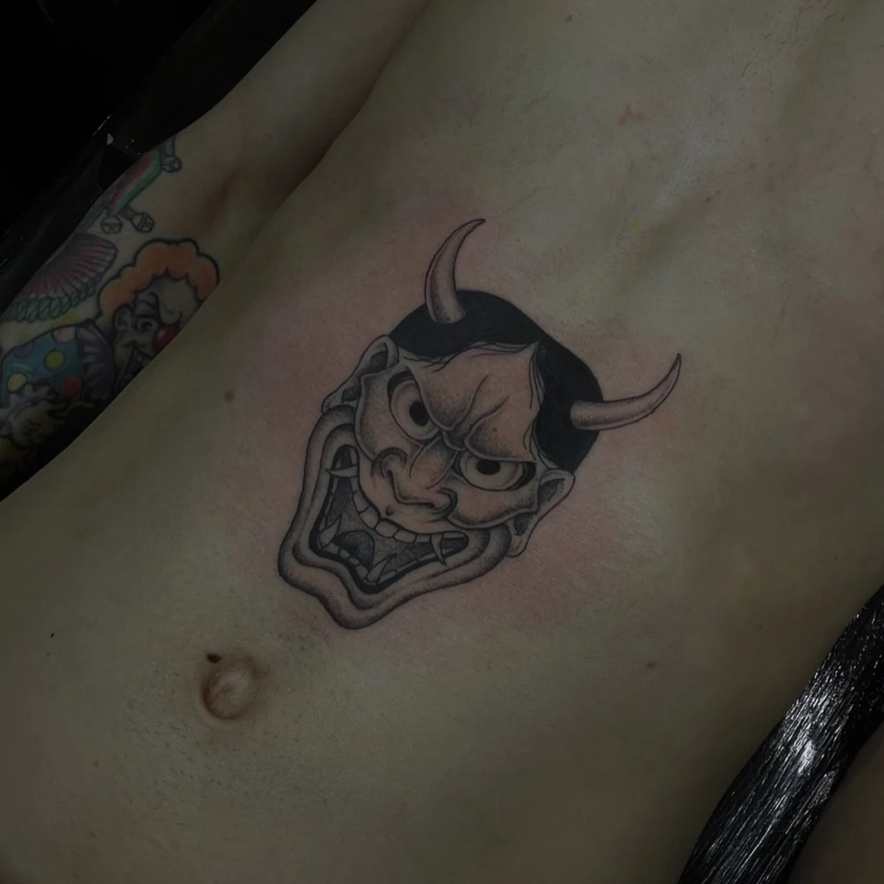Tattoo of a fierce, grinning demon face with horns on a person's skin.