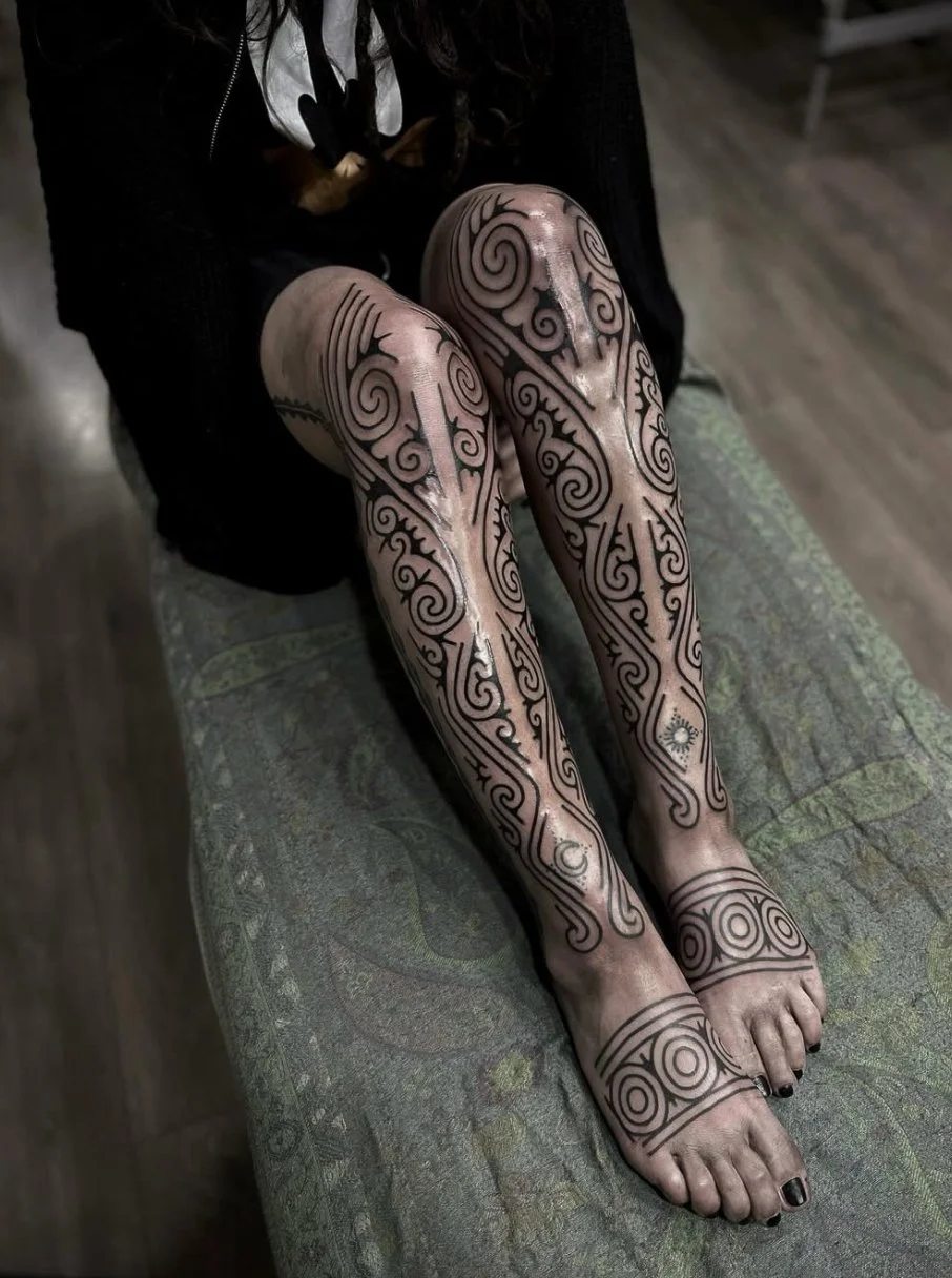 Person with tattooed legs sitting on a bench, showing black and gray tribal-style tattoos covering from thighs to toes.