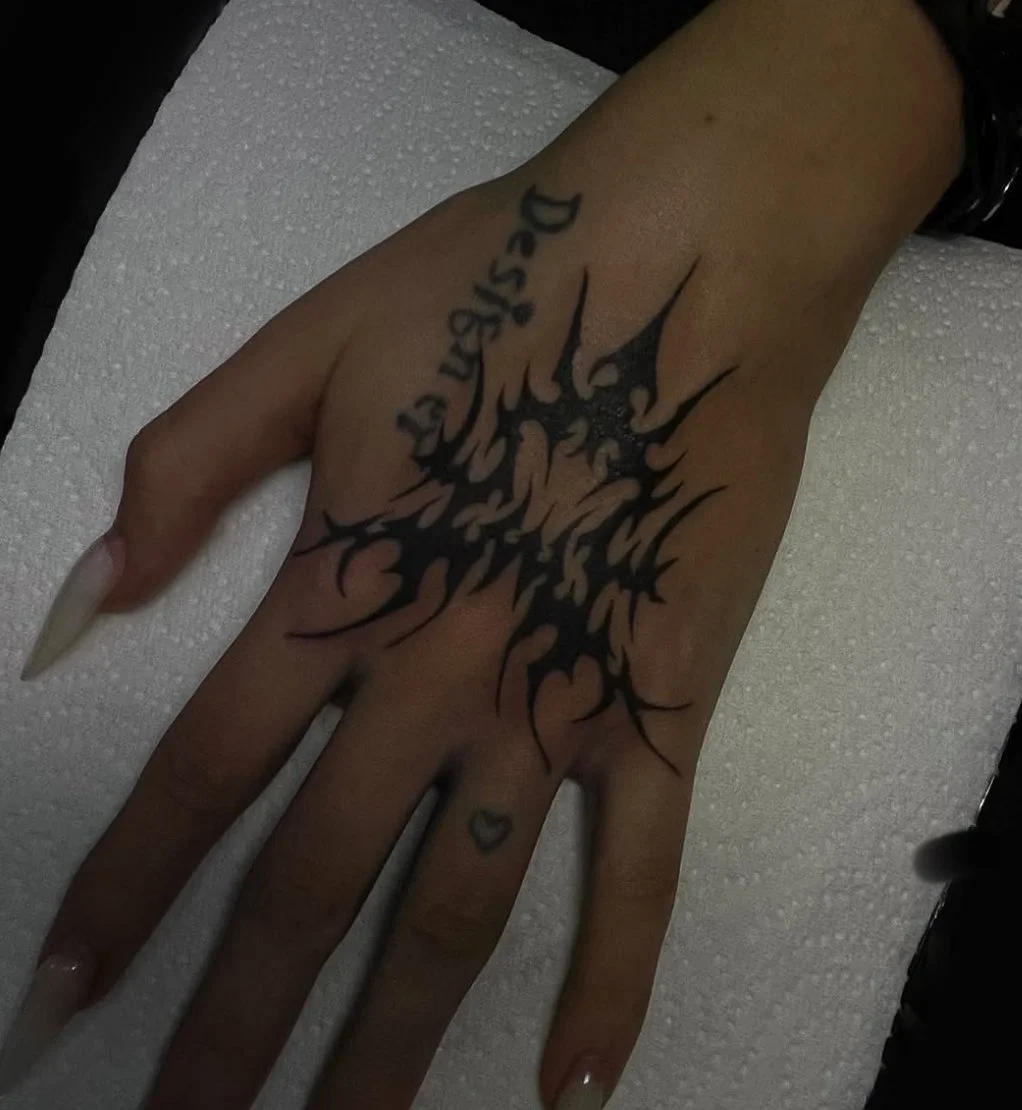Close-up of a hand with a black tribal-style tattoo covering the back of the hand and extending onto the fingers. The tattoo features sharp, jagged lines and a small heart tattoo on the middle finger. The hand is resting on a white textured surface.